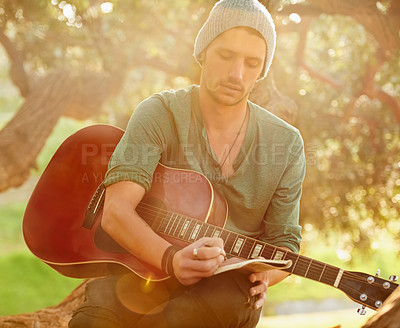 Buy stock photo Shot of a handsome young man sitting in a park with a guitar writing song on a notepad