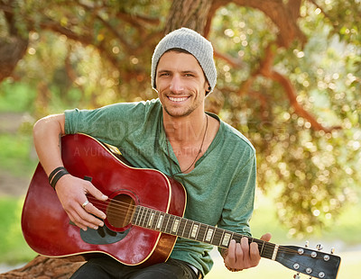 Buy stock photo Portrait of a handsome young man sitting outside in a park playing guitar