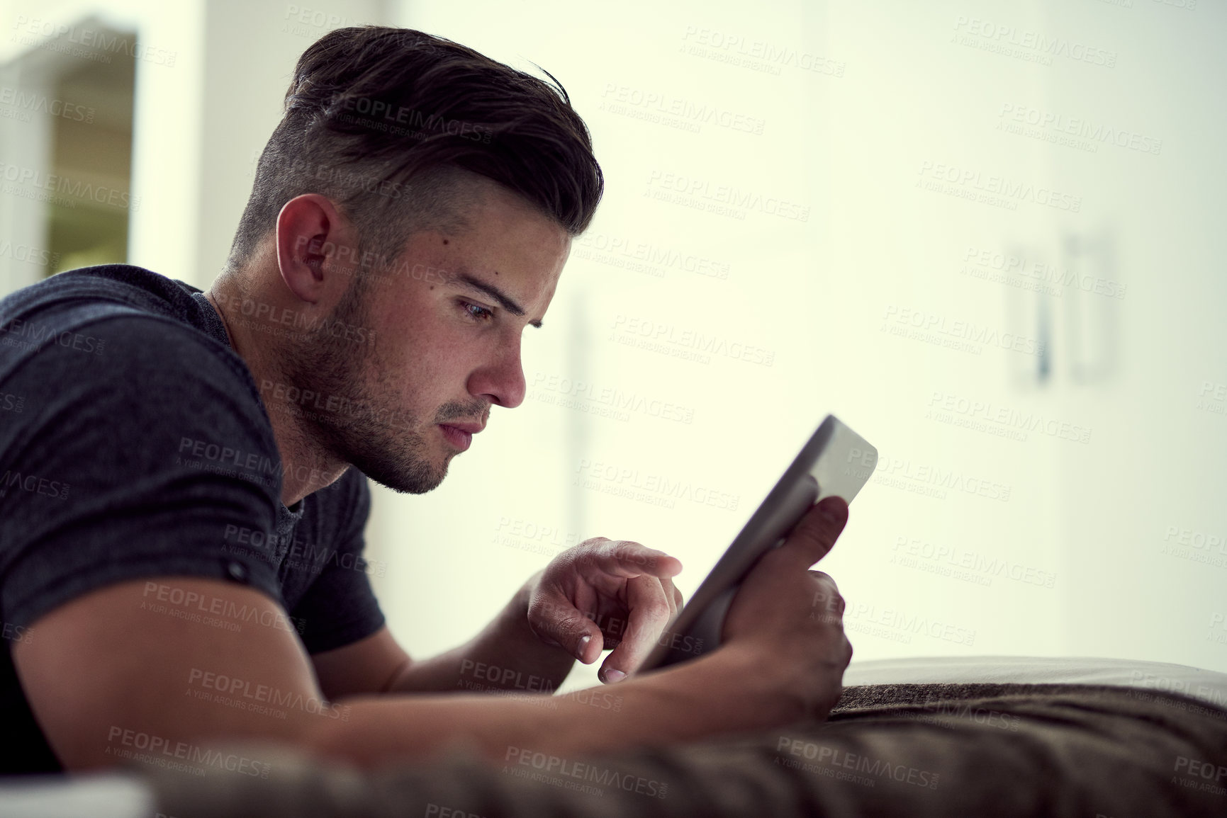 Buy stock photo Shot of a young man using his wireless tablet while lying on his bed at home