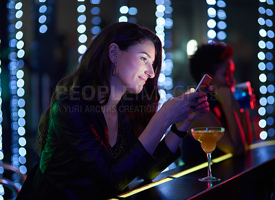 Buy stock photo Shot of a smiling young woman reading a text message while sitting at the bar in a nightclub