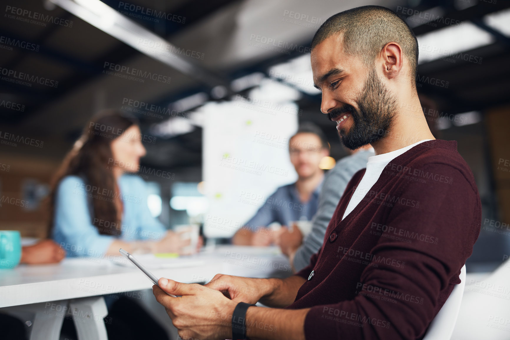 Buy stock photo Shot of a young man sitting at a table in an office using a digital tablet with colleagues working in the background