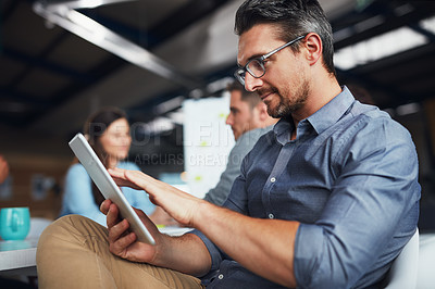 Buy stock photo Shot of a man sitting at a table in an office using a digital tablet with colleagues working in the background