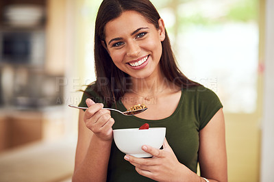 Buy stock photo Portrait of a happy young woman eating a bowl of muesli while standing in her kitchen