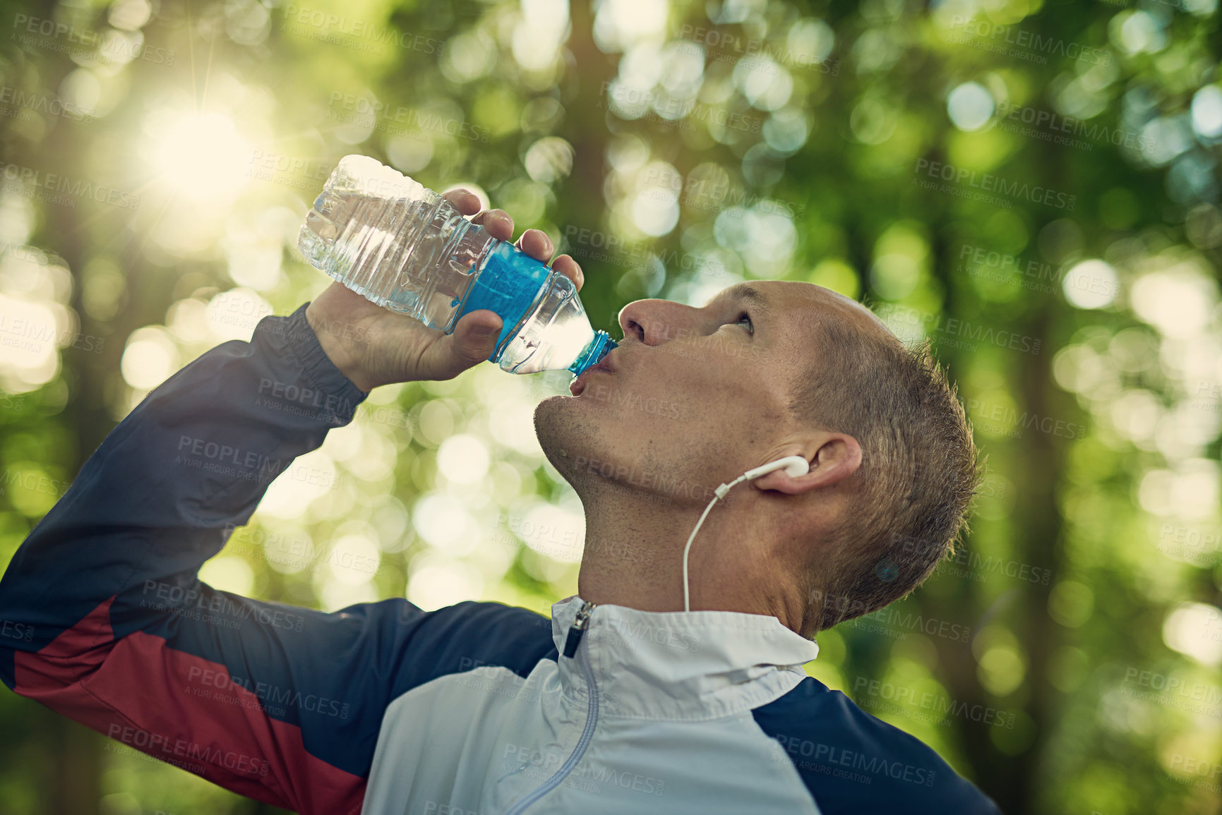 Buy stock photo Running, fitness or man drinking water in park with earphones for music, wellness or training motivation in nature. Sport, liquid or thirsty runner with aqua drink in forest streaming workout podcast