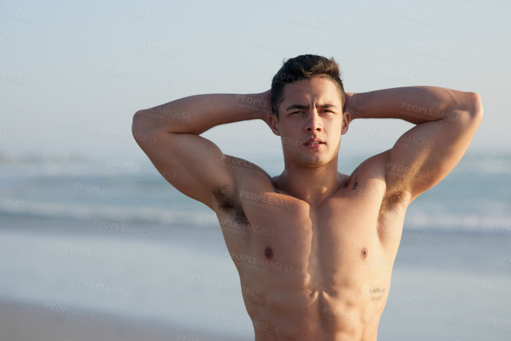 Buy stock photo Cropped portrait of a handsome young man standing with his hands behind his head at the beach