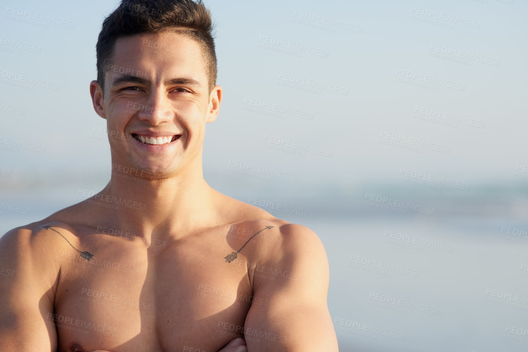 Buy stock photo Cropped portrait of a handsome young man standing with his arms folded at the beach