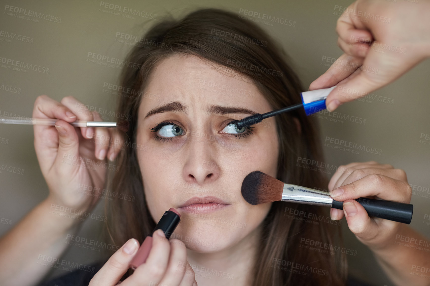 Buy stock photo Concept shot of a nervous looking young woman with an assortment of brushes applying makeup to her face