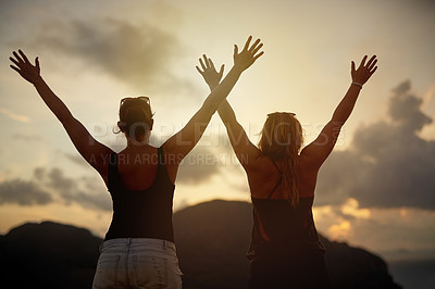 Buy stock photo Rearview shot of two young friends with their arms raised silhouetted against a sunset