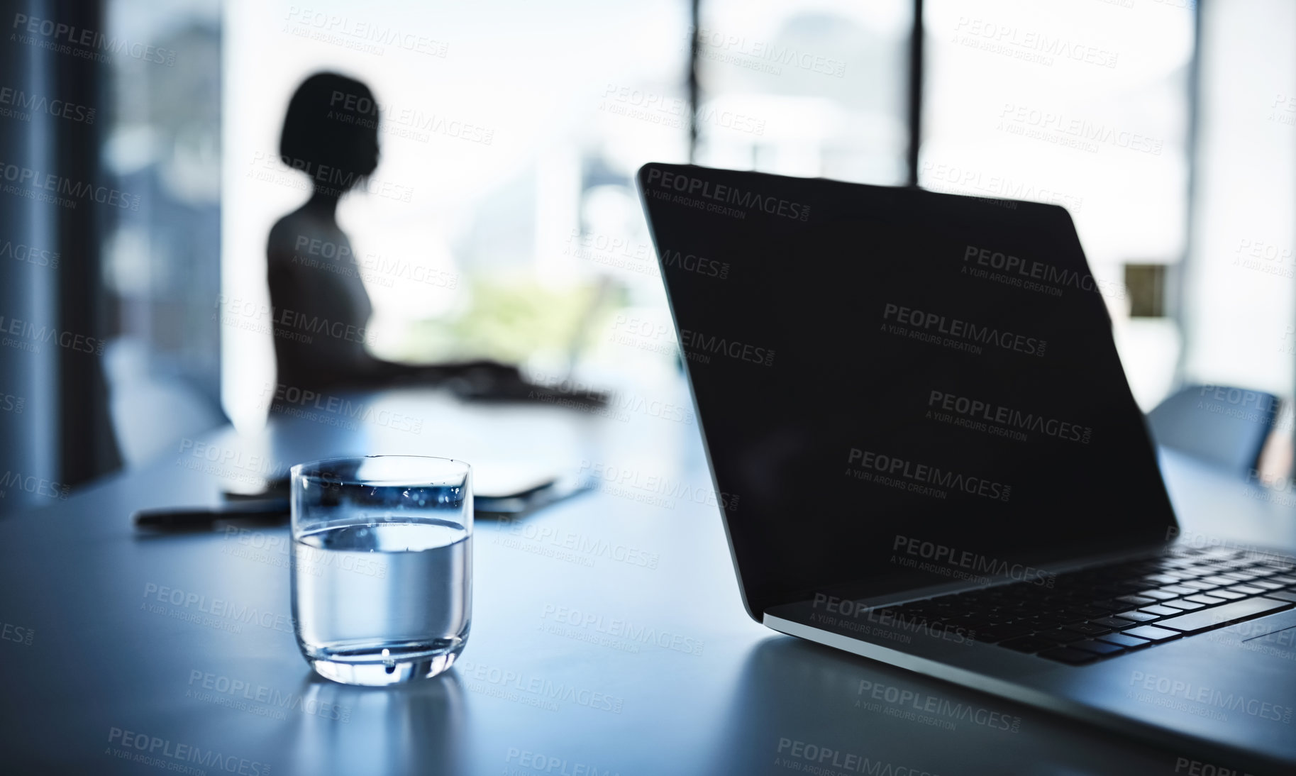 Buy stock photo Cropped shot of a laptop and a glass of water on a boardroom table with a businesswoman in the background