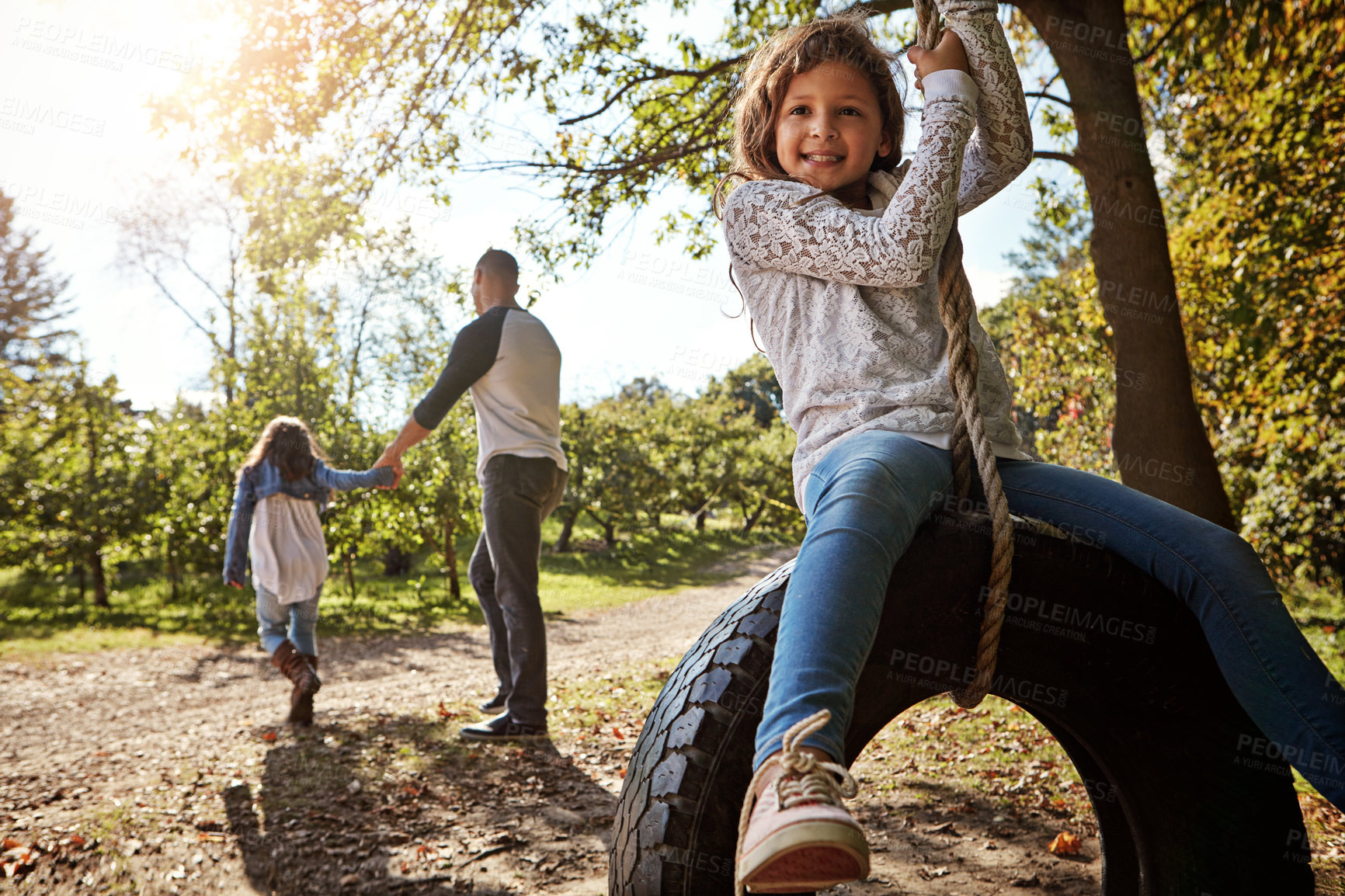 Buy stock photo Portrait of a happy little girl swinging on a tyre with her father and sister in the background