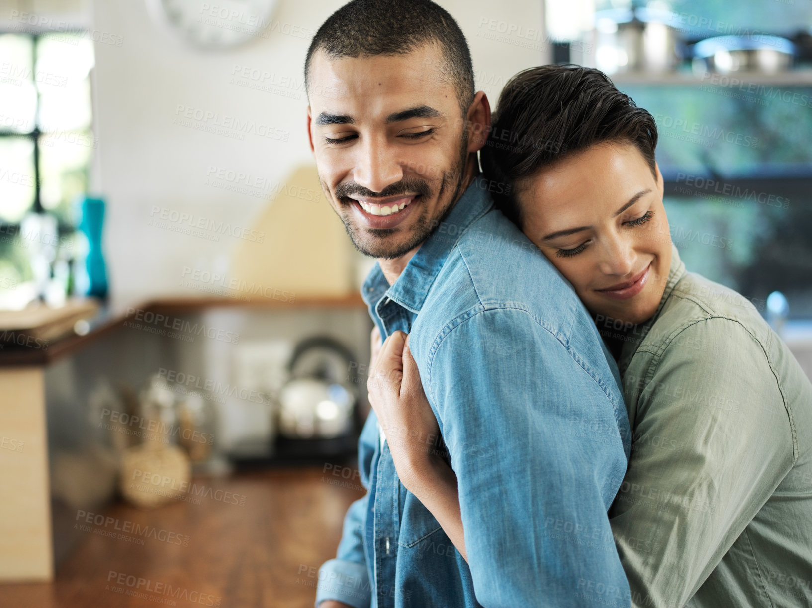 Buy stock photo Shot of a young woman embracing her husband from behind while standing in their kitchen