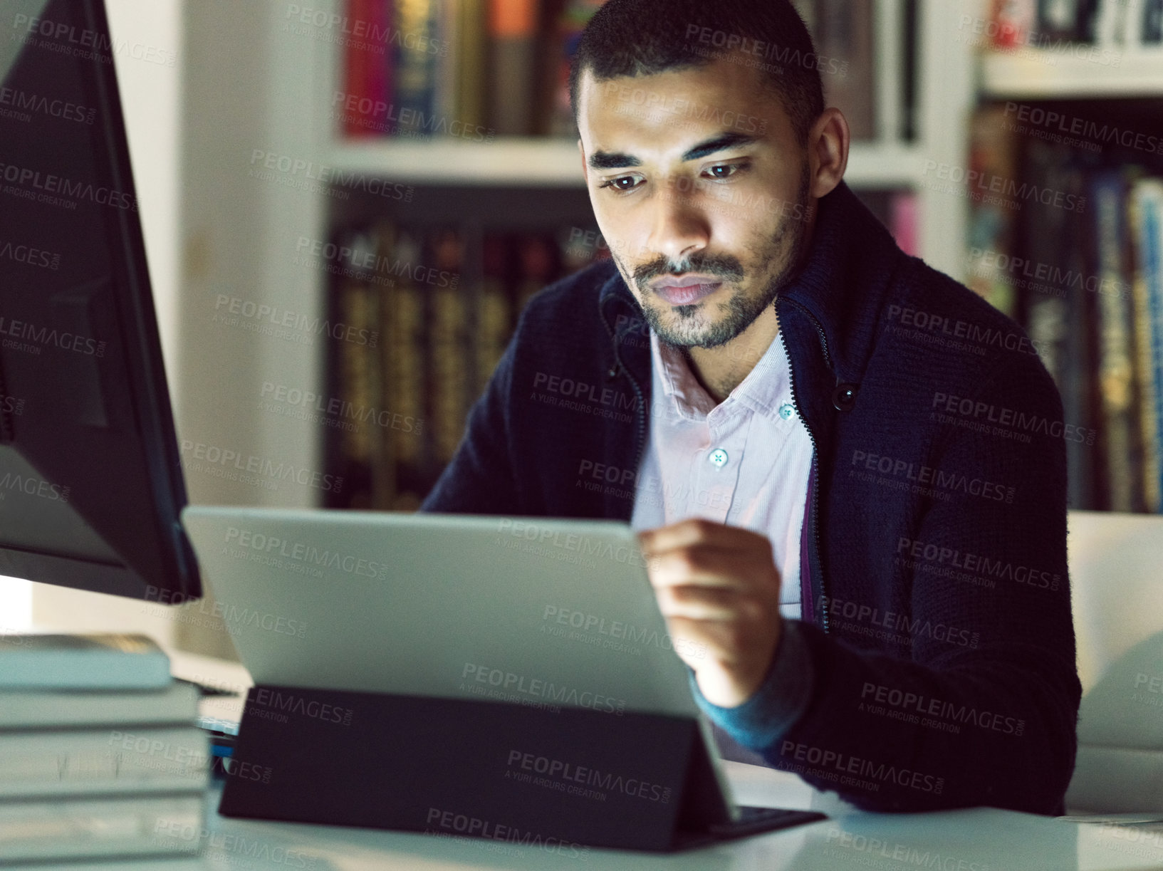 Buy stock photo Shot of a focused young man working on a digital tablet in his home office in the early evening