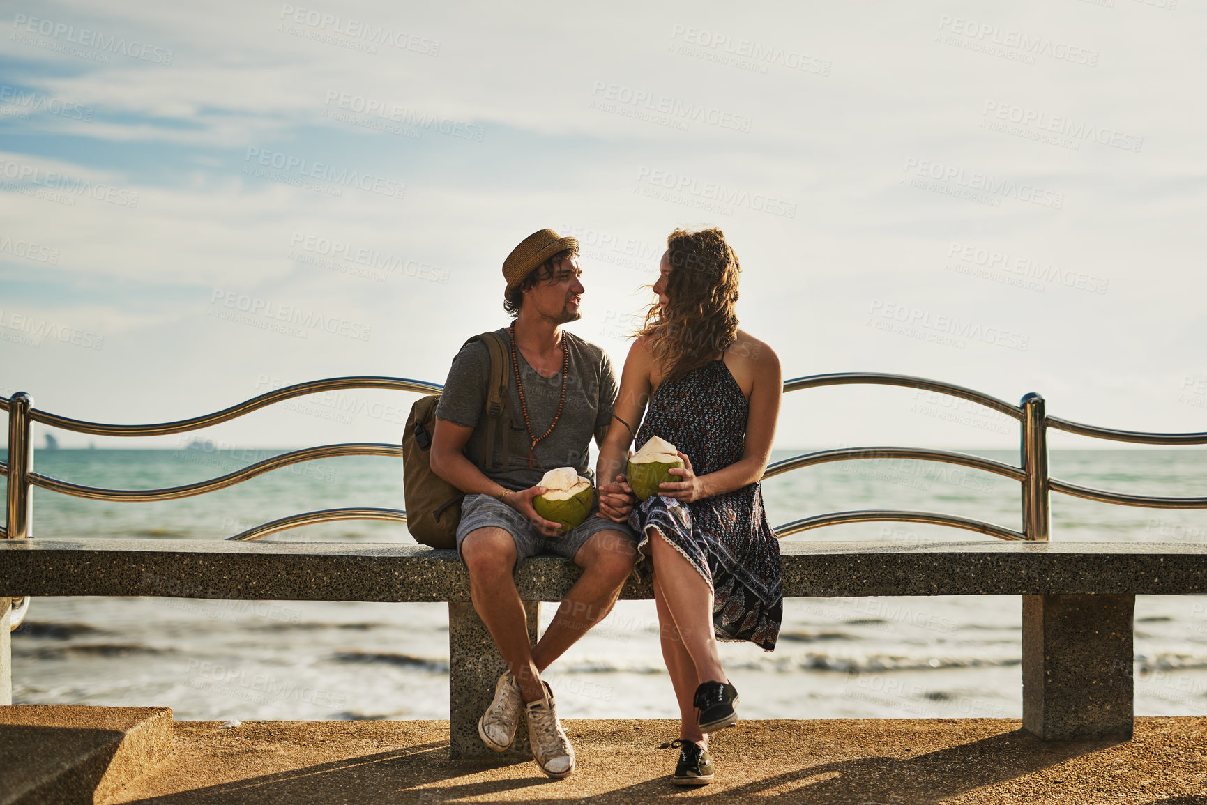 Buy stock photo Shot of a happy young couple enjoying cocktails while relaxing on a bench by the beach