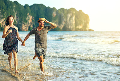 Buy stock photo Shot of a happy young couple holding hands while running along the beach