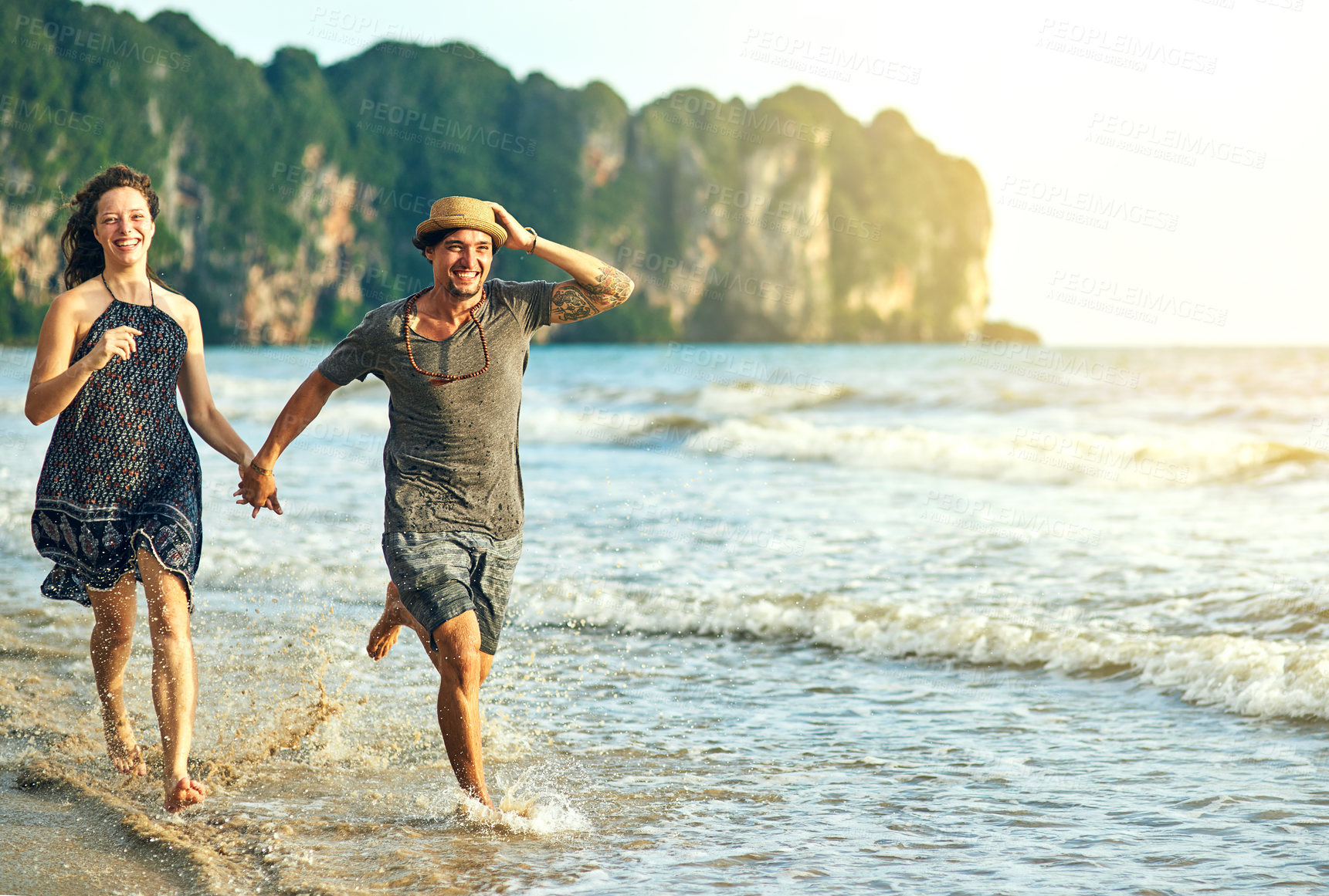 Buy stock photo Shot of a happy young couple holding hands while running along the beach