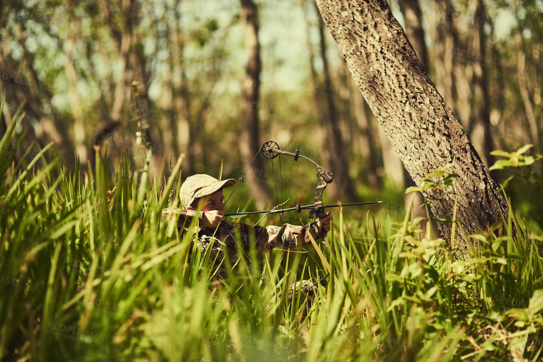 Buy stock photo Shot of a cute little boy in camouflage playing with his bow and arrow in the woods