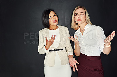 Buy stock photo Portrait of two young businesswomen shrugging against a dark background