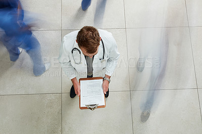 Buy stock photo High angle shot of a male doctor looking at a patient's file in a busy hospital