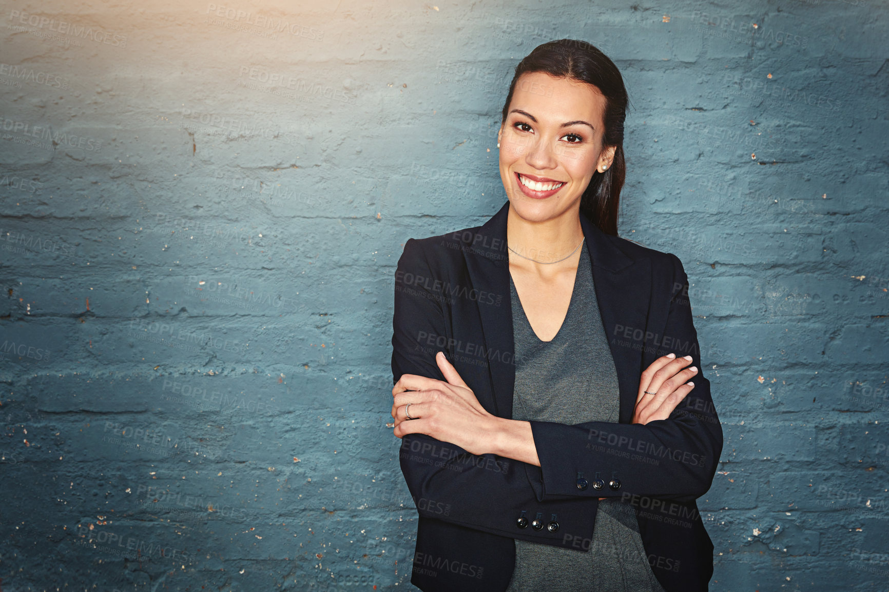 Buy stock photo Portrait of a confident young businesswoman posing against a brick wall with her arms crossed