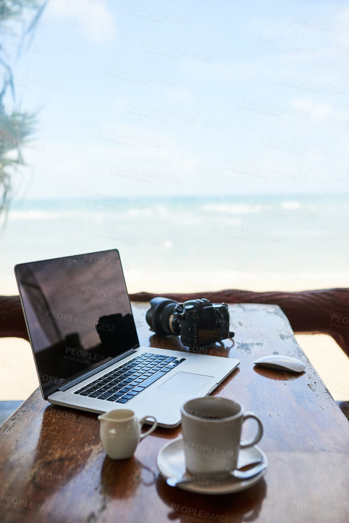 Buy stock photo Shot of a laptop and a cup of coffee on a table with a view of the beach in the background