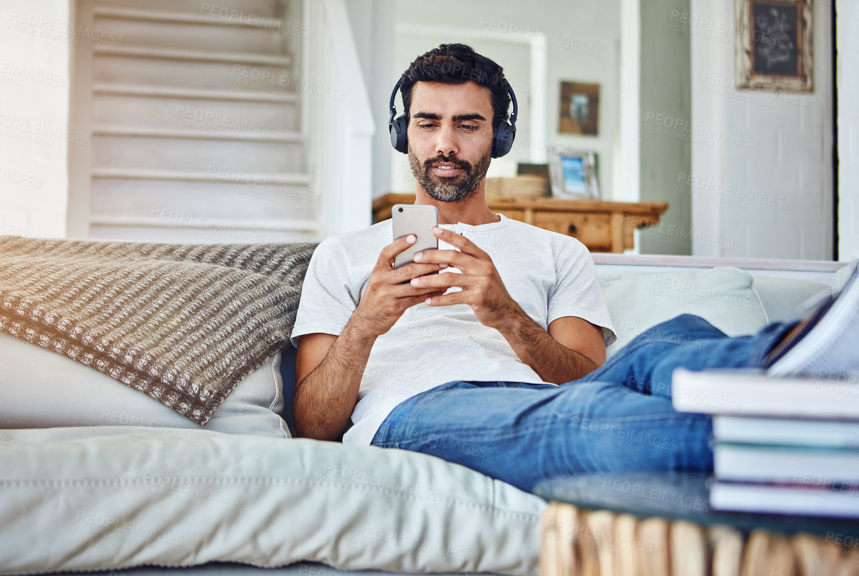 Buy stock photo Man, phone and video with headphones on sofa for sound, audio and streaming service in living room with relax. Person, smartphone and mobile app for listening to music, radio and subscription in home