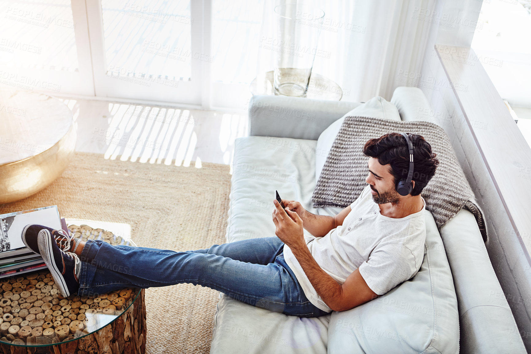 Buy stock photo House, man and smartphone with headphones on sofa to listen to music, radio and network at home. Living room, male person and mobile app for streaming podcast, online games or social media to relax
