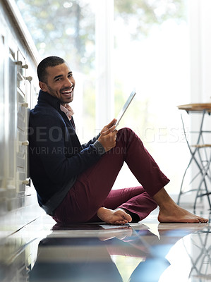 Buy stock photo Portrait of a smiling young man sitting on his kitchen floor using a digital tablet