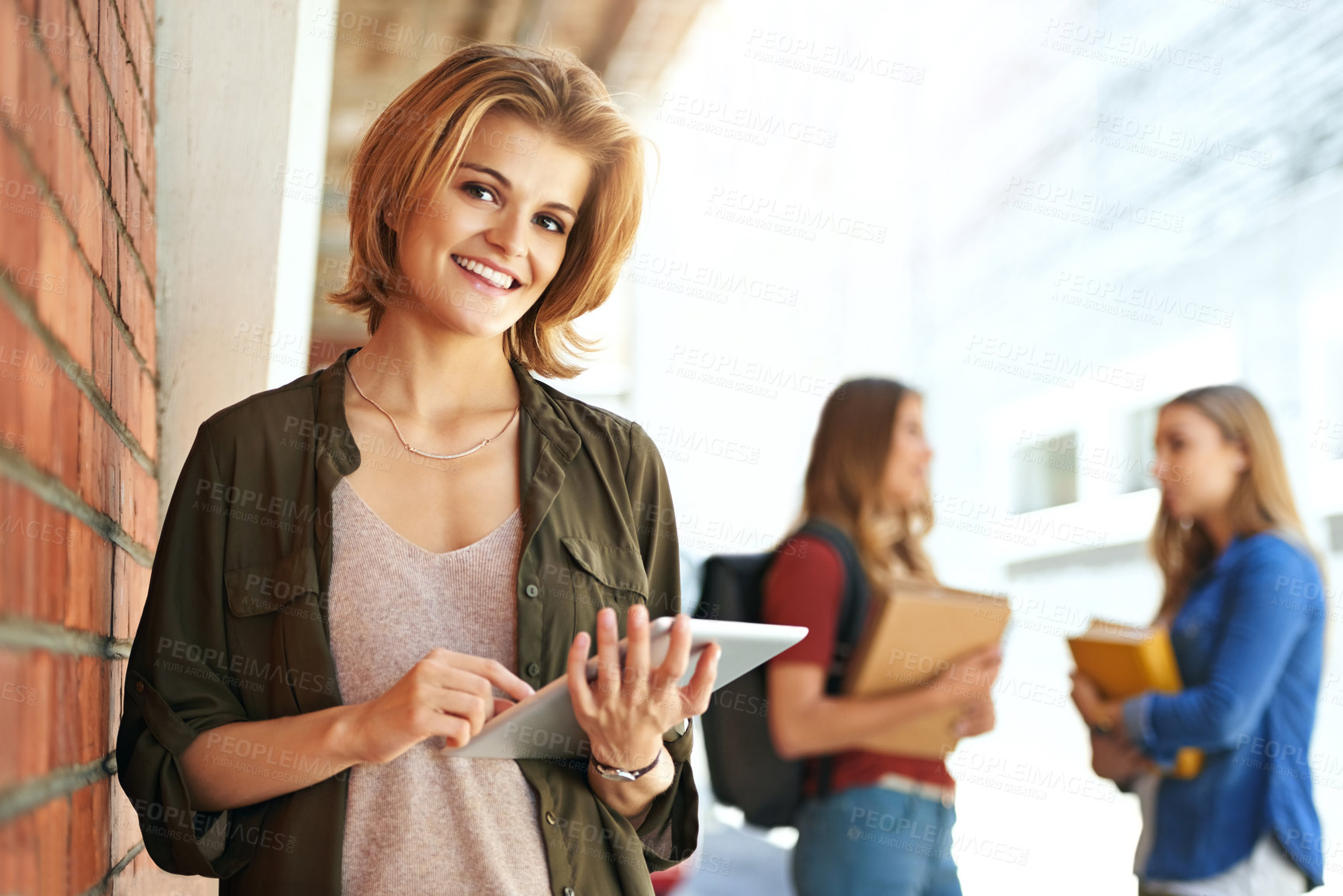 Buy stock photo Portrait of a smiling female university student standing on campus using a digital tablet with friends in the background