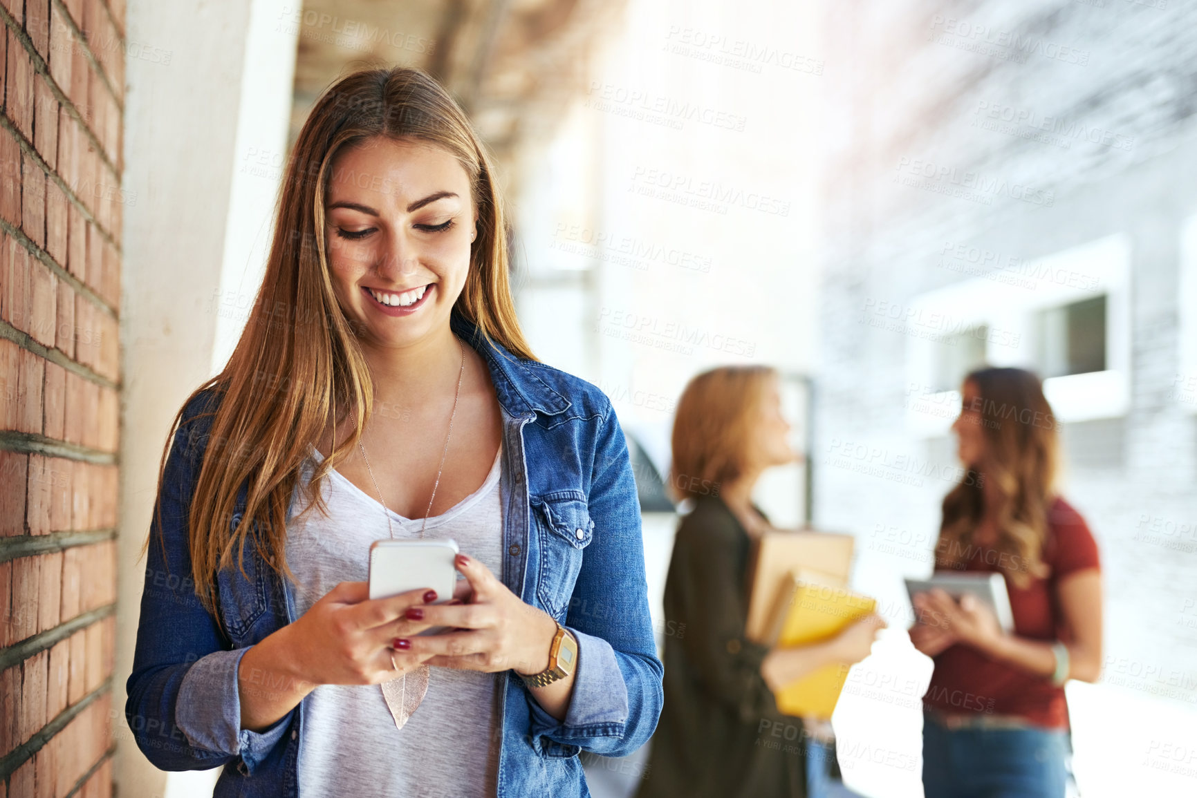 Buy stock photo Shot of a smiling female university student standing on campus using a cellphone with friends in the background