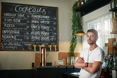Buy stock photo Portrait of a serious young bartender posing with his arms crossed behind the bar