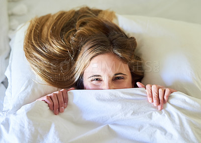 Buy stock photo Portrait of a playful young woman hiding under the covers in her bed