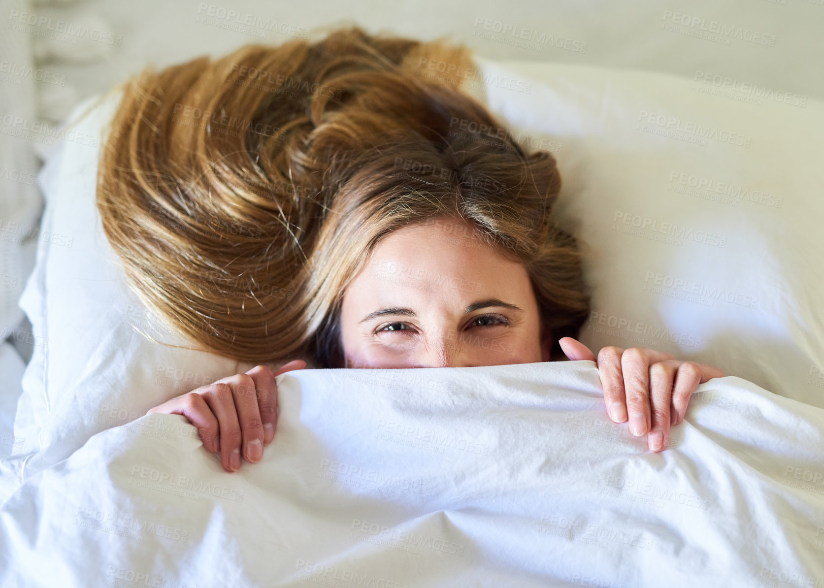 Buy stock photo Portrait of a playful young woman hiding under the covers in her bed
