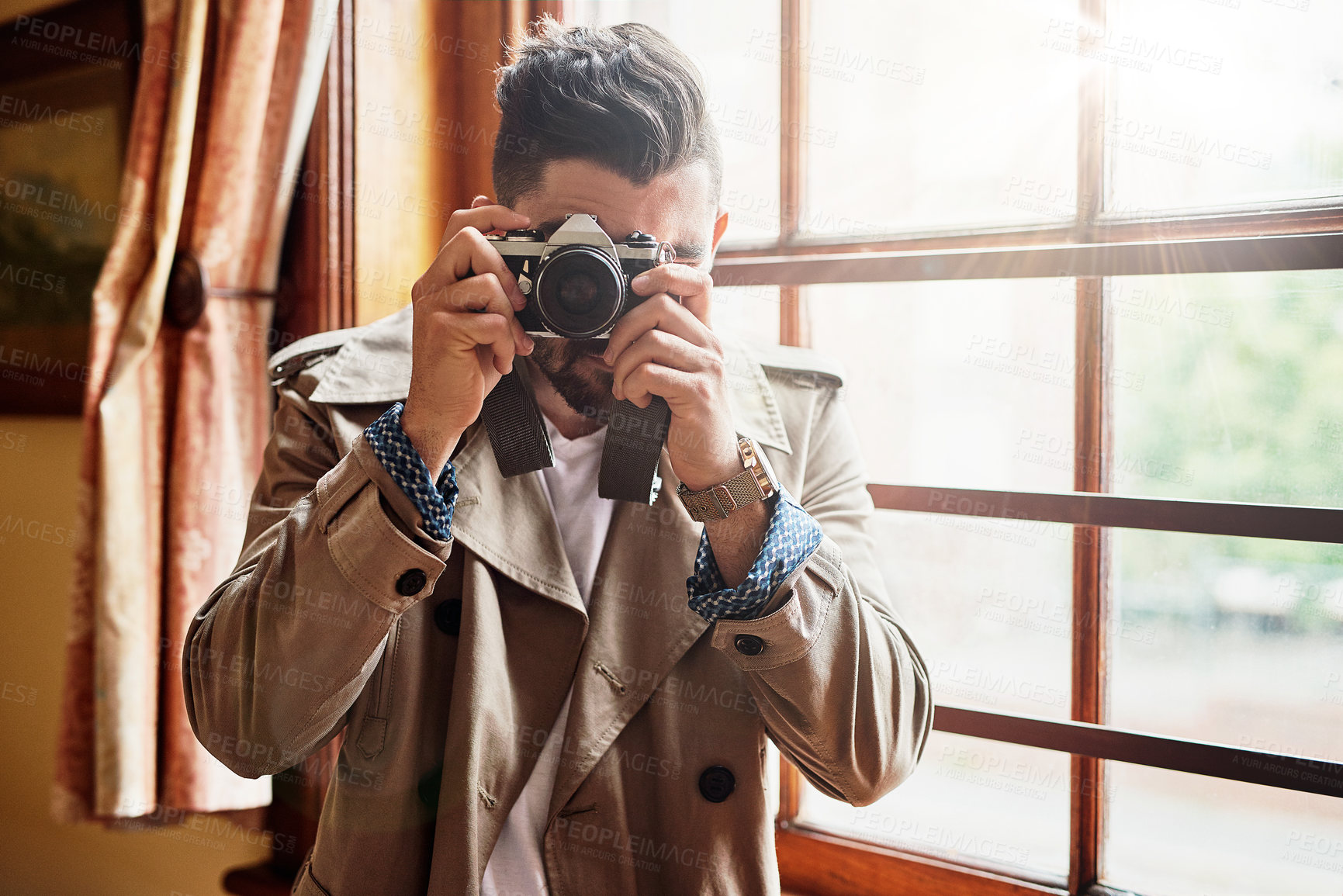 Buy stock photo Shot of a handsome young man  standing by a window at home using a camera