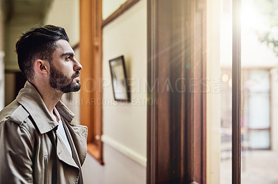 Buy stock photo Shot of a handsome young man standing by a window looking deep in thought