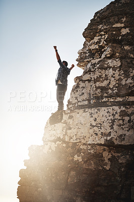 Buy stock photo Shot of a young man standing on a mountain cliff with his arms raised