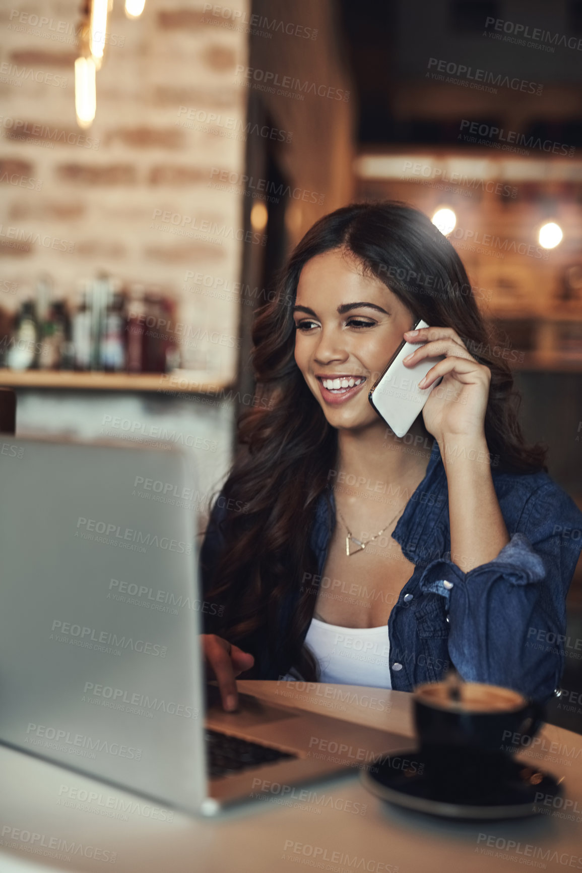 Buy stock photo Shot of a young woman answering her cellphone while using her laptop in a coffee shop