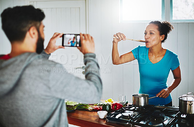 Buy stock photo Shot of a man filming his wife on his smartphone while she cooks in their kitchen at home