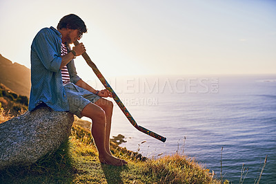 Buy stock photo Shot of a young man playing the didgeridoo while sitting at a viewpoint outside