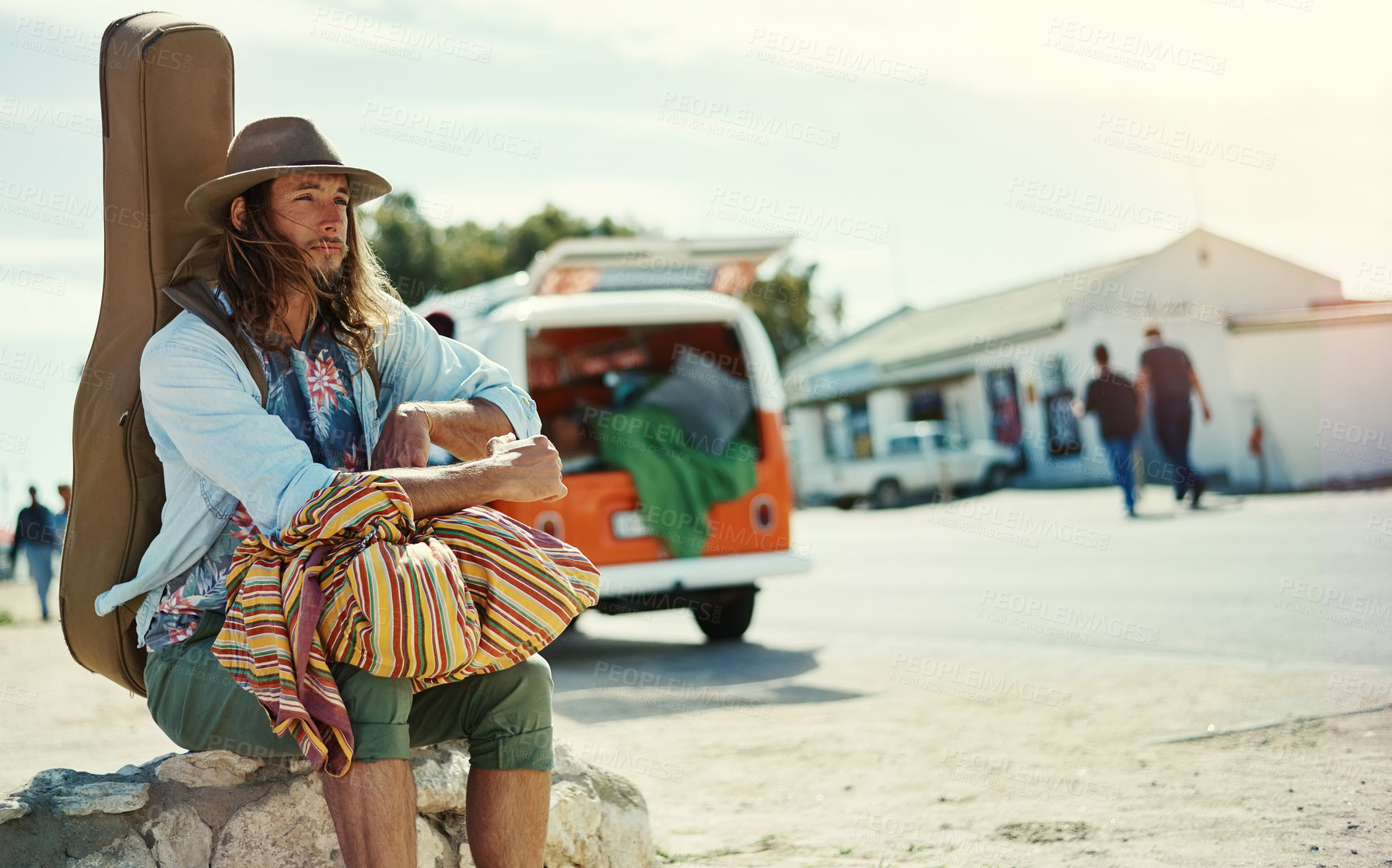 Buy stock photo Shot of a young man with a guitar on his back sitting at the side of the road with broken down van in the background