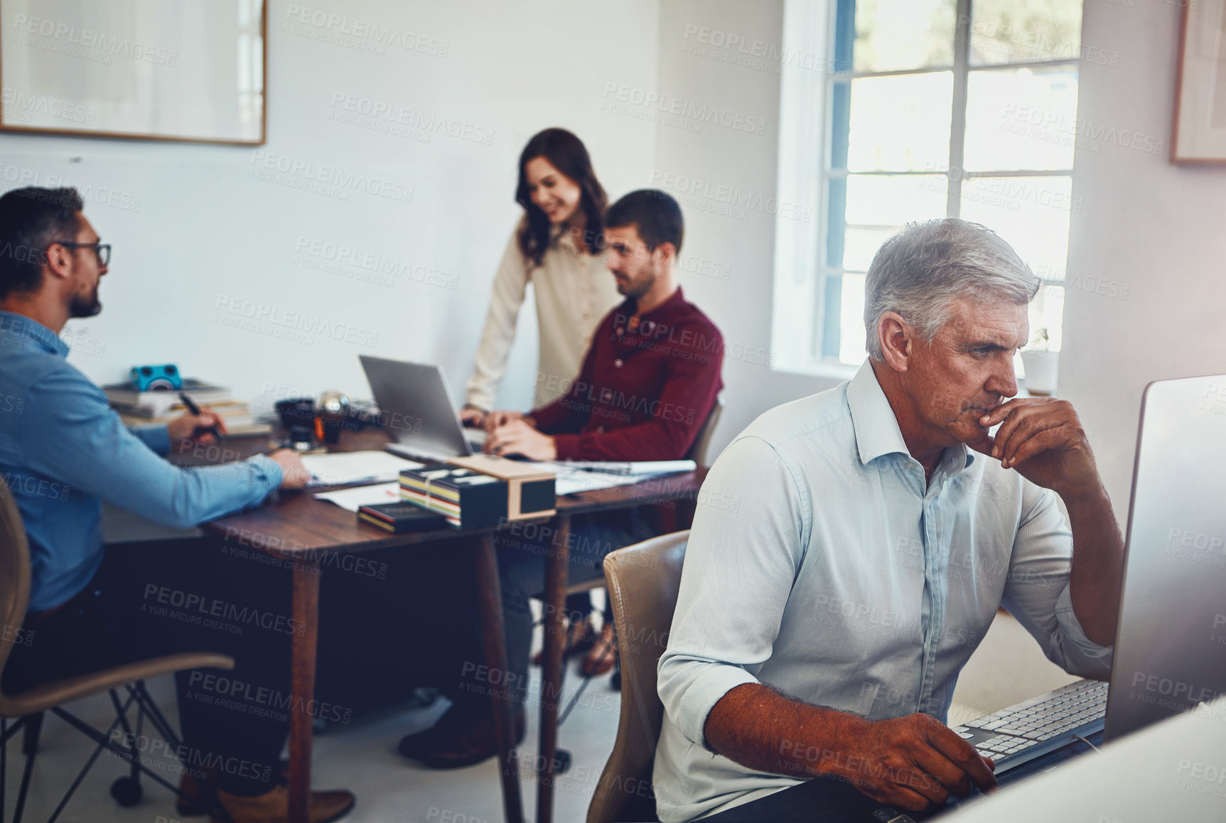 Buy stock photo Shot of a mature man working in the office with his colleagues in the background