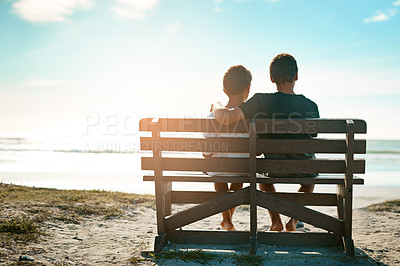Buy stock photo Rearview shot of two unidentifiable brothers sitting on a bench together by the beach