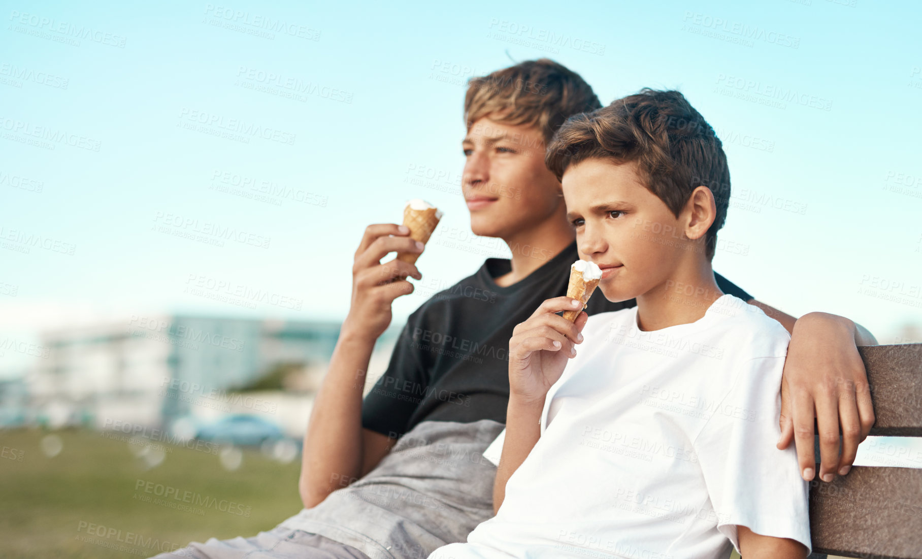 Buy stock photo Shot of two happy brothers eating ice-cream cones while sitting on a bench by the beach