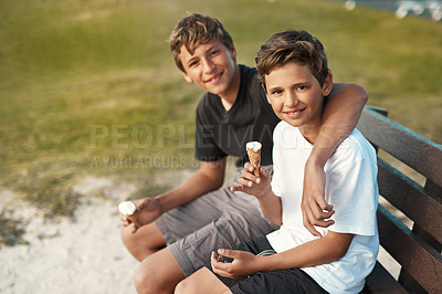 Buy stock photo Portrait of two happy brothers eating ice-cream cones while sitting on a bench by the beach
