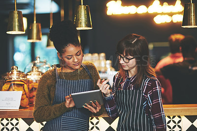 Buy stock photo Shot of two young women using a digital tablet together while working at a coffee shop