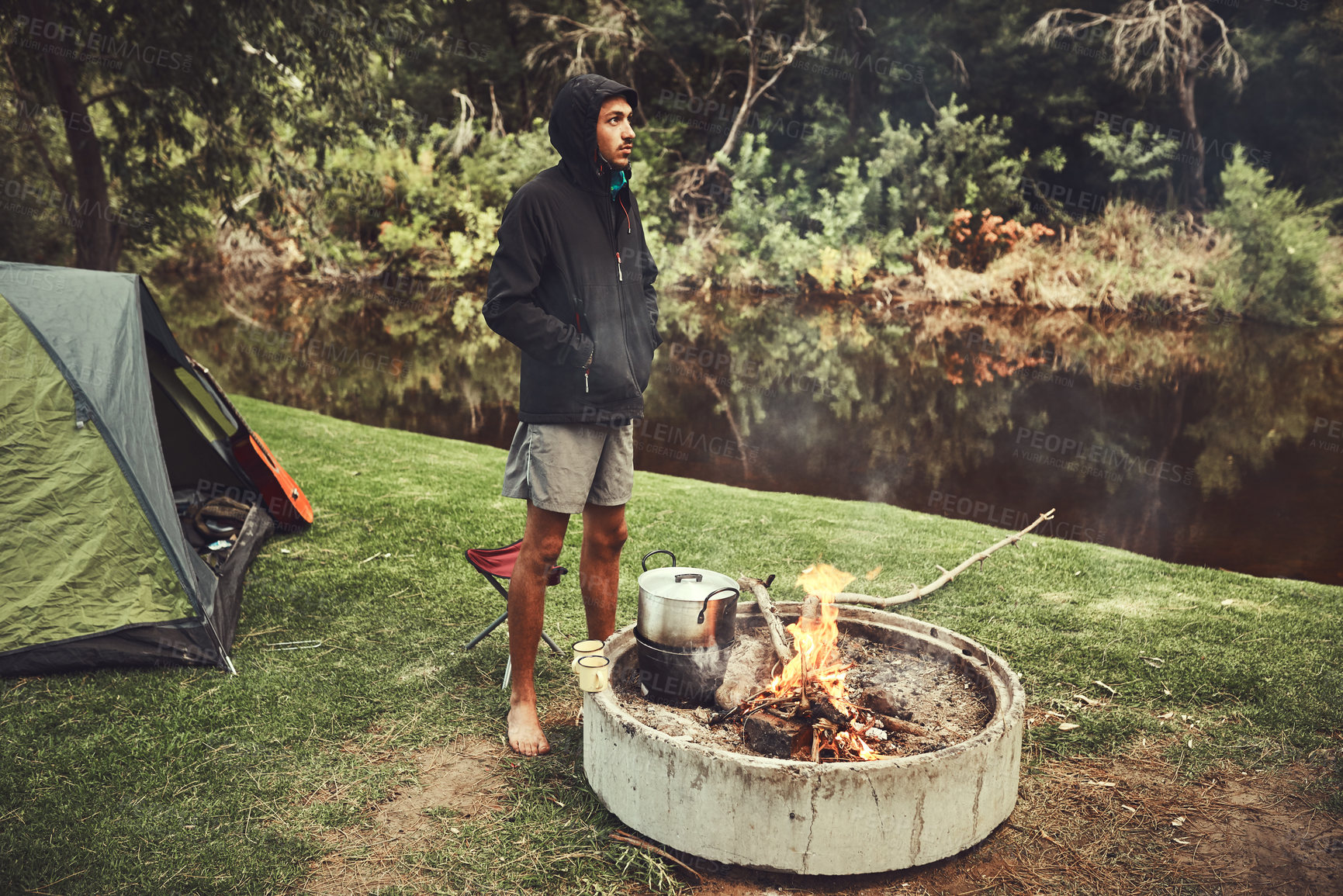 Buy stock photo Camping, thinking and man in woods with fire for adventure, travel and vacation by lake. Wellness, tent and male person in nature for eating, nutrition and reflection of trees in water in forest