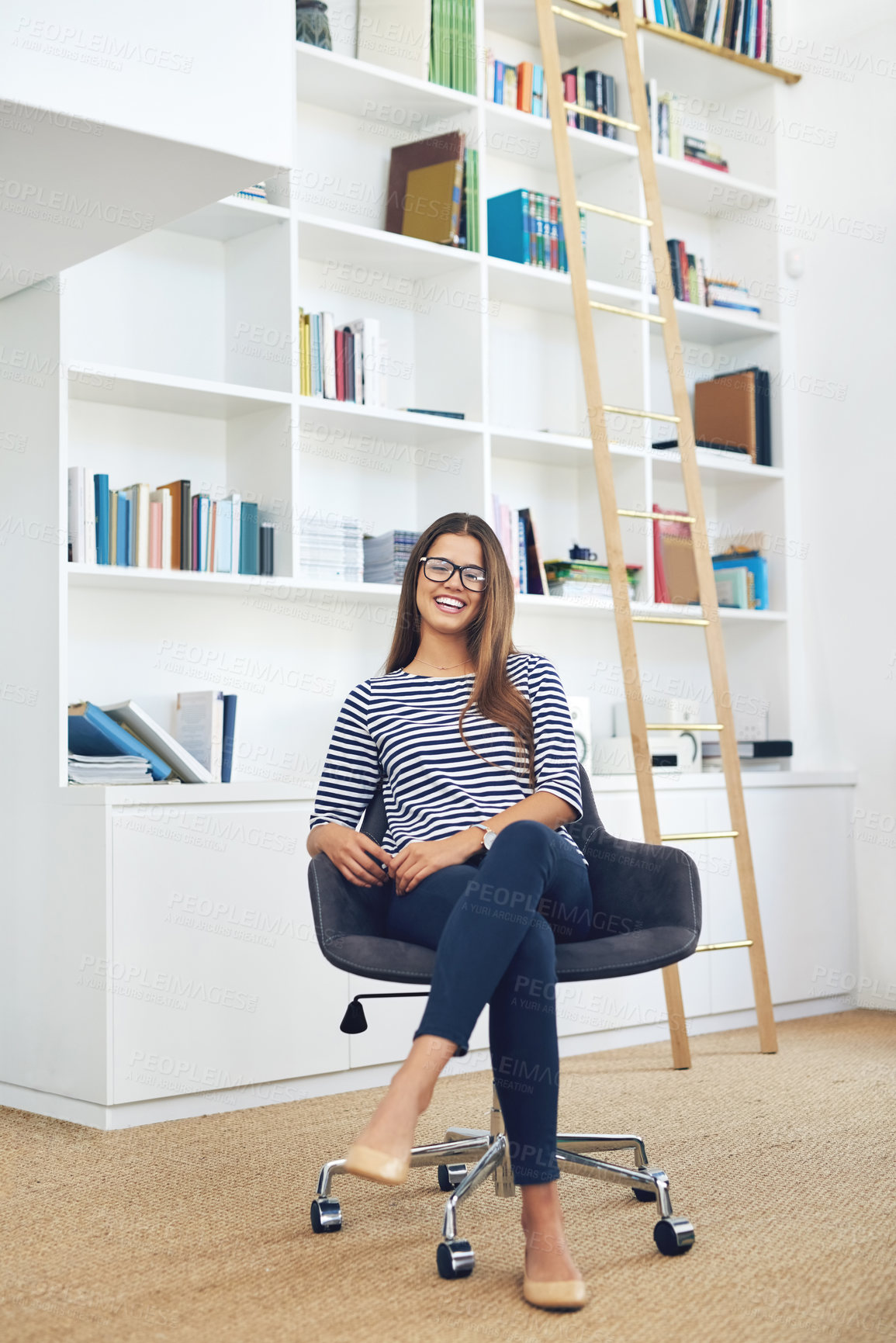 Buy stock photo Portrait of a smiling young woman wearing glasses sitting in front of bookshelves at home