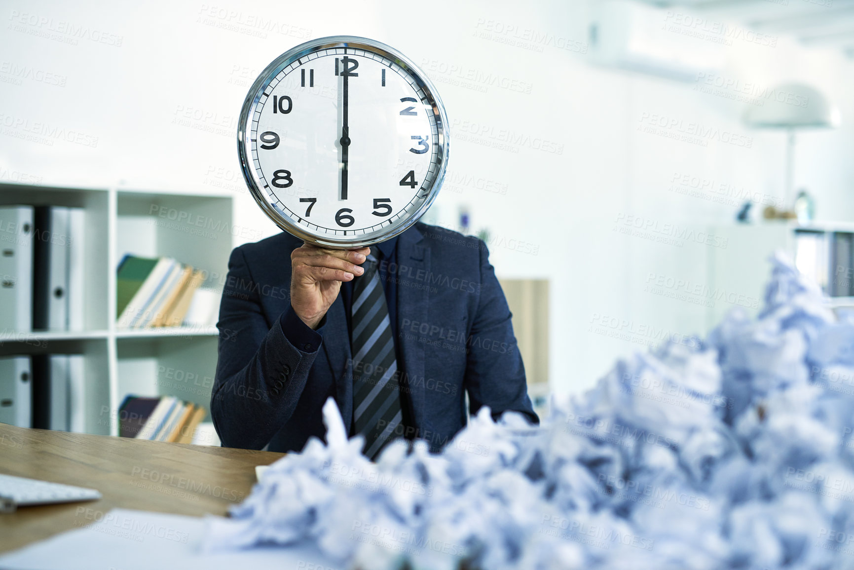 Buy stock photo Shot of a businessman sitting at his desk overwhelmed by paperwork holding up a clock in front of his face