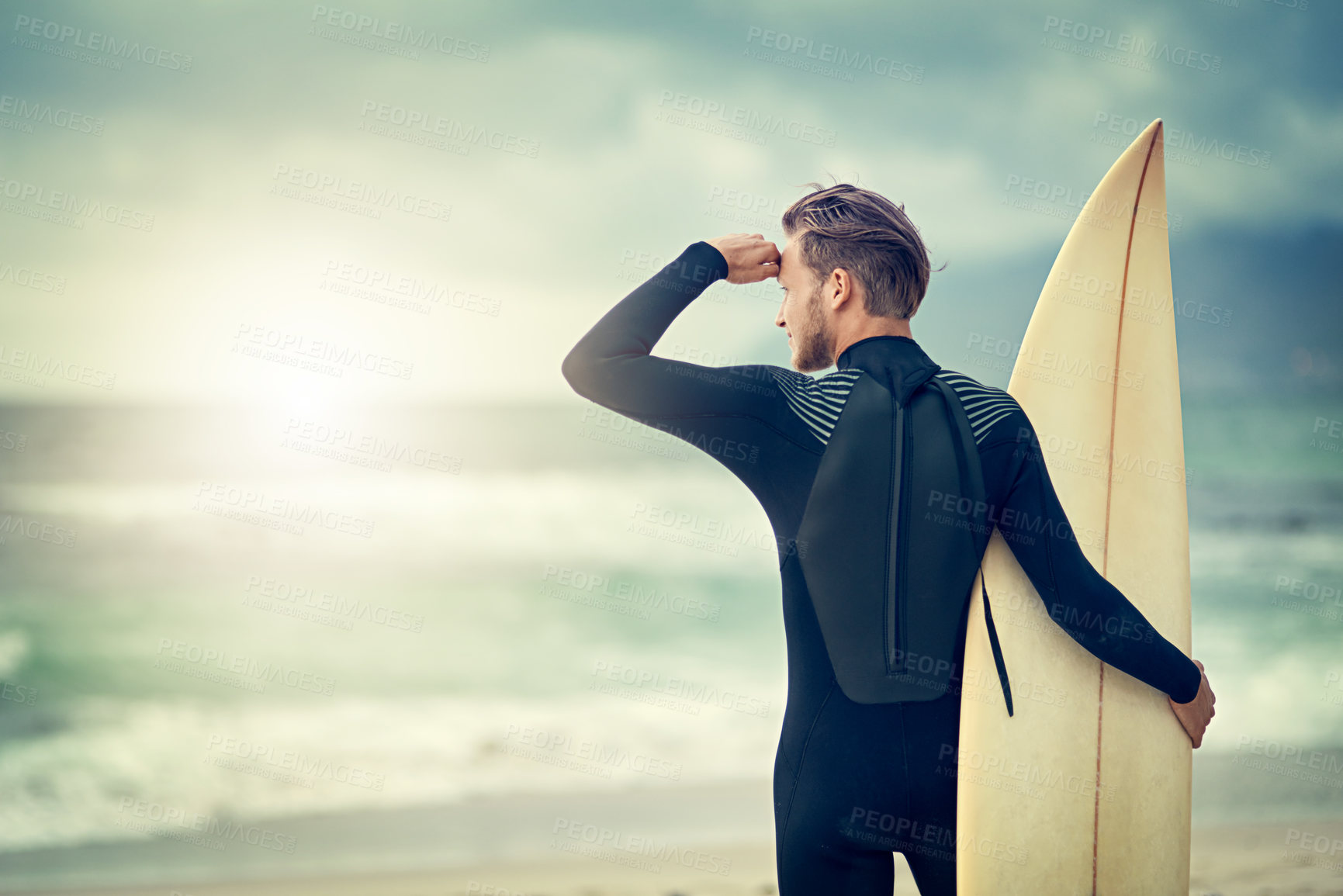 Buy stock photo Shot of a laid-back young surfer watching the waves while holding his surfboard at the beach