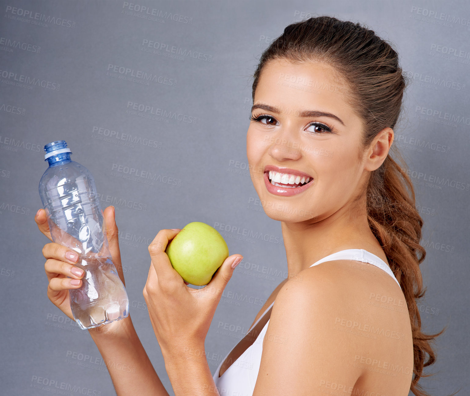 Buy stock photo Studio portrait of an attractive young woman holding an apple and a bottle of water against a grey background