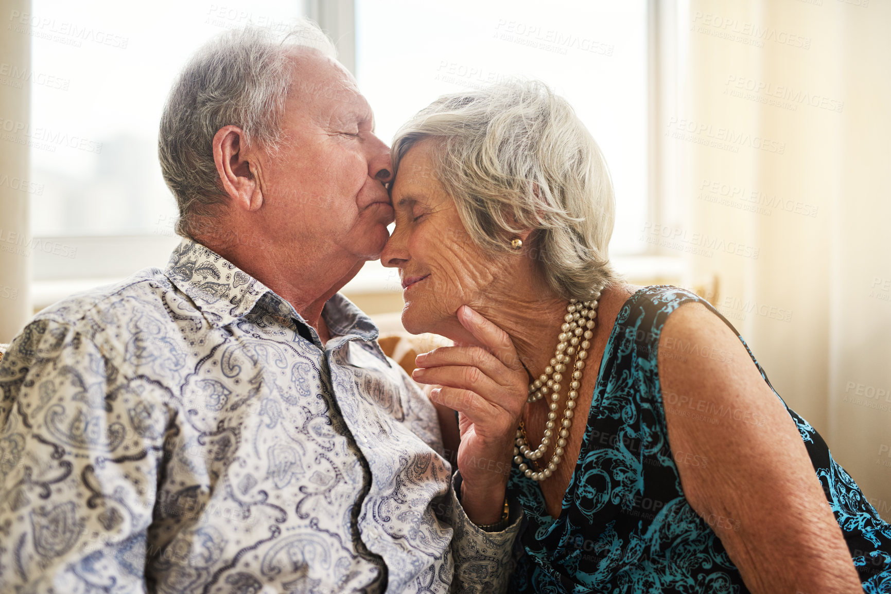 Buy stock photo Senior, couple and kiss at nursing home with love, forehead and bonding in living room. Retirement, care and relax in a house happy with elderly people on a sofa with marriage and support together